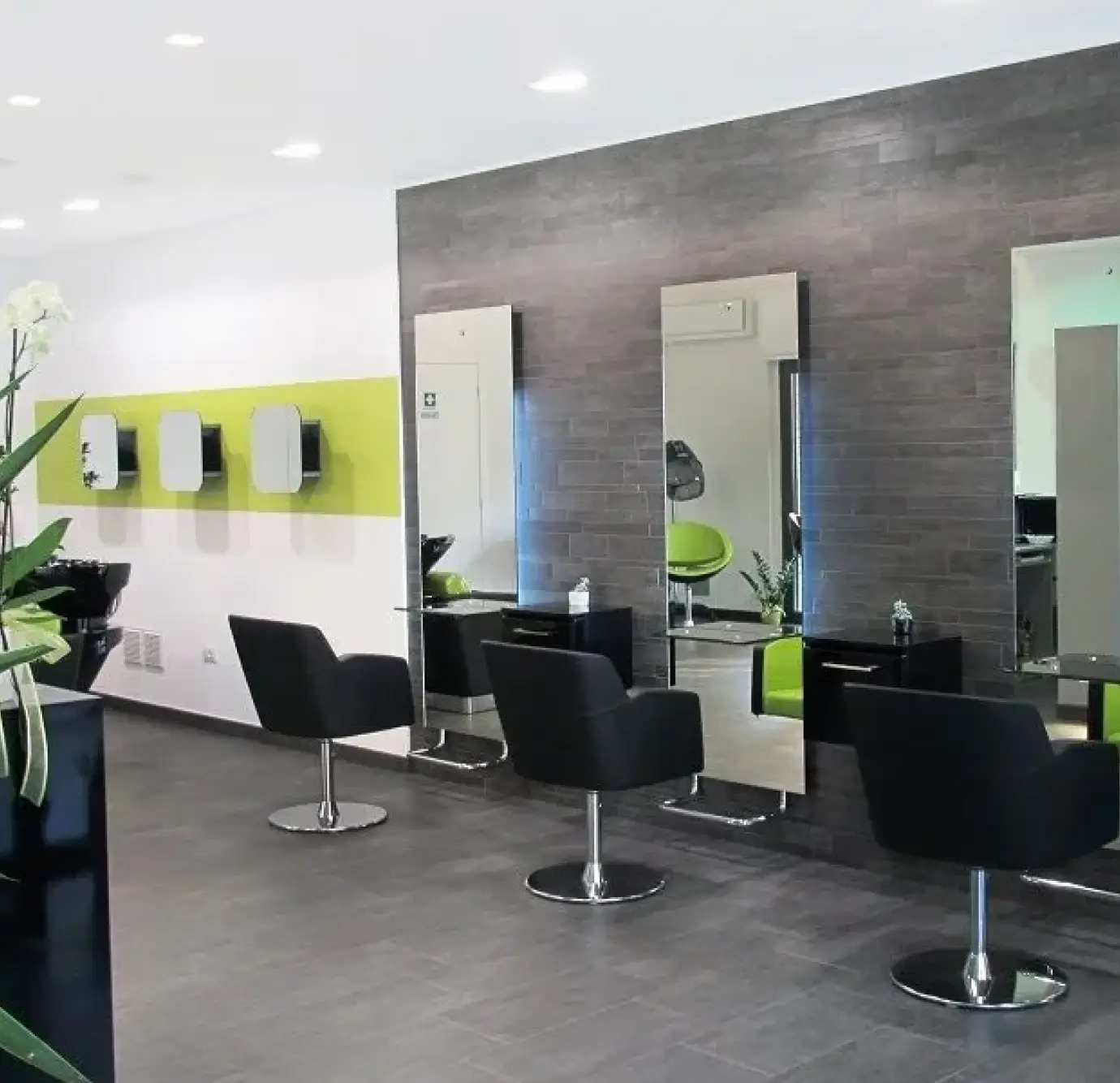 Modern hair salon interior with black styling chairs, tall rectangular mirrors on a dark gray textured wall, green accent stripe, and recessed lighting