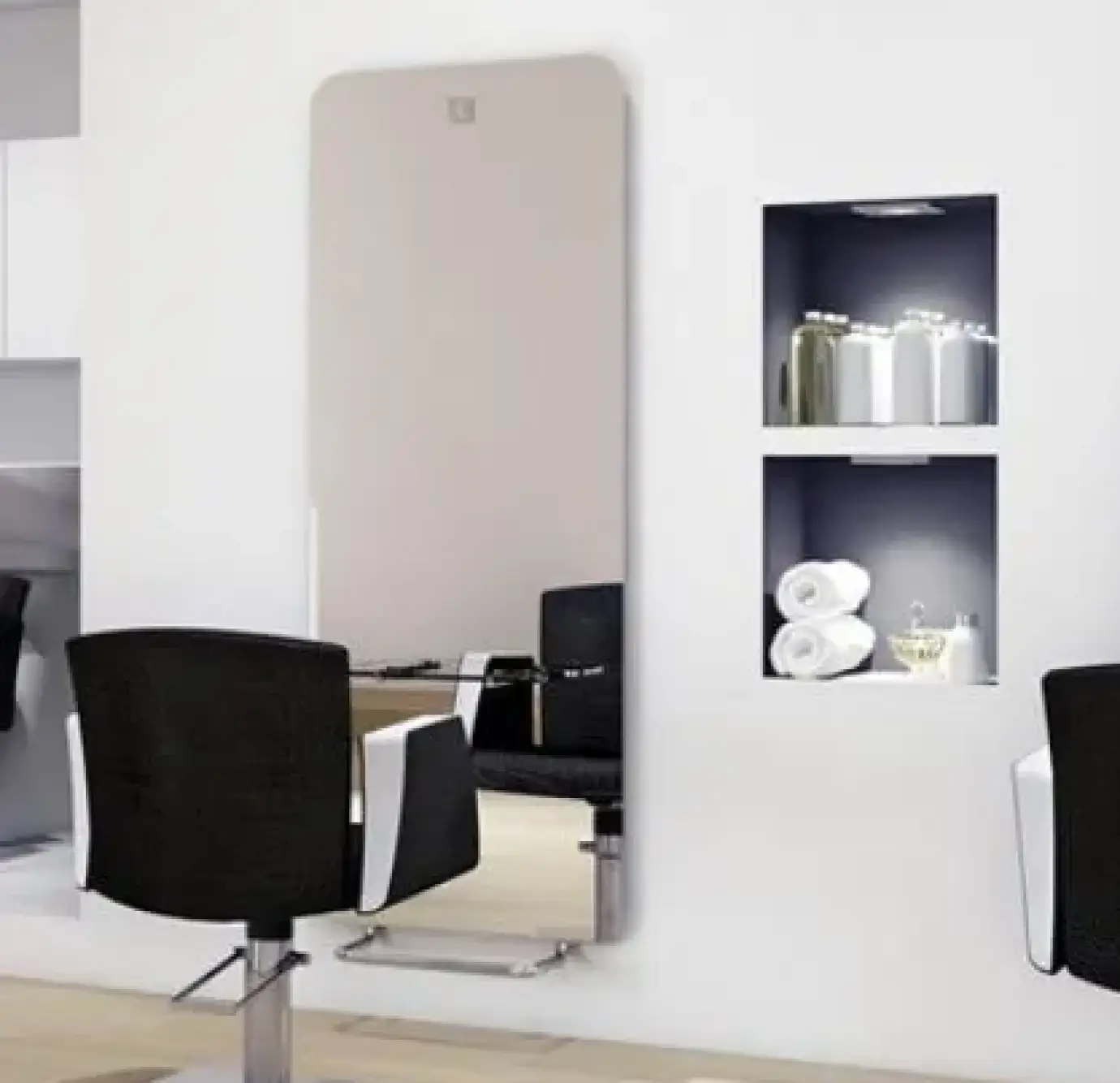 Minimalist salon styling station with a large frameless wall mirror, black styling chair, and two illuminated wall niches displaying folded towels and hair care products