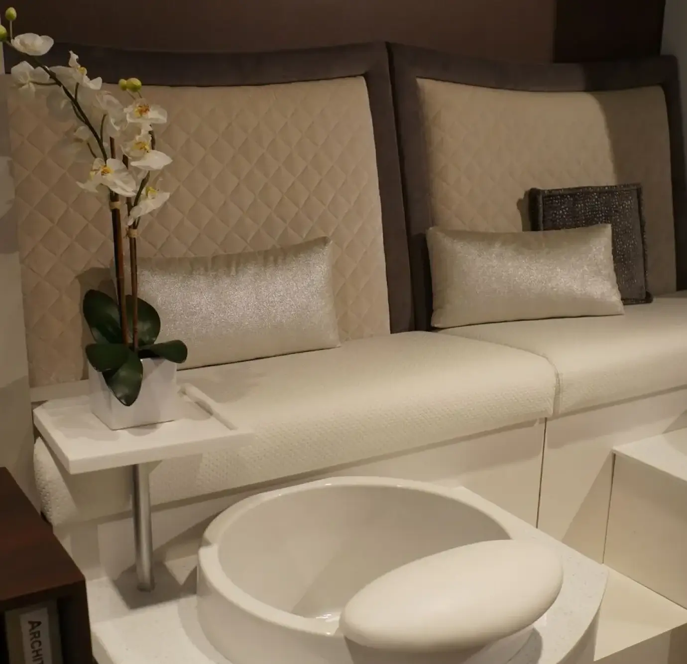 Luxury nail salon pedicure stations with cream quilted high-back seating, silver shimmer lumbar pillows, white orchid centerpiece, and white porcelain foot basins