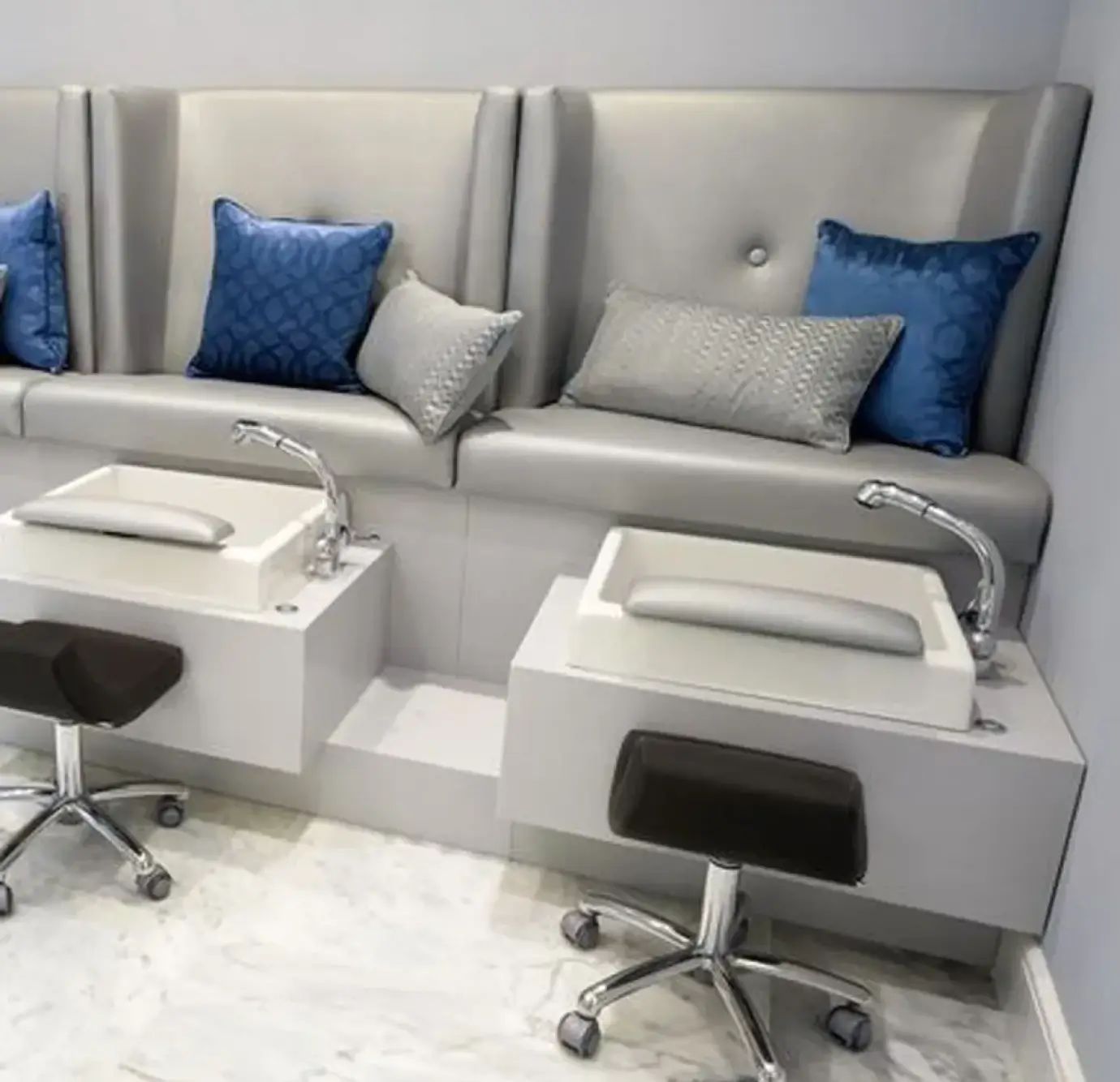 Modern nail salon pedicure bench with silver faux leather high-back seating, blue and grey decorative pillows, white foot basins with chrome faucets, and rolling technician stools on marble flooring