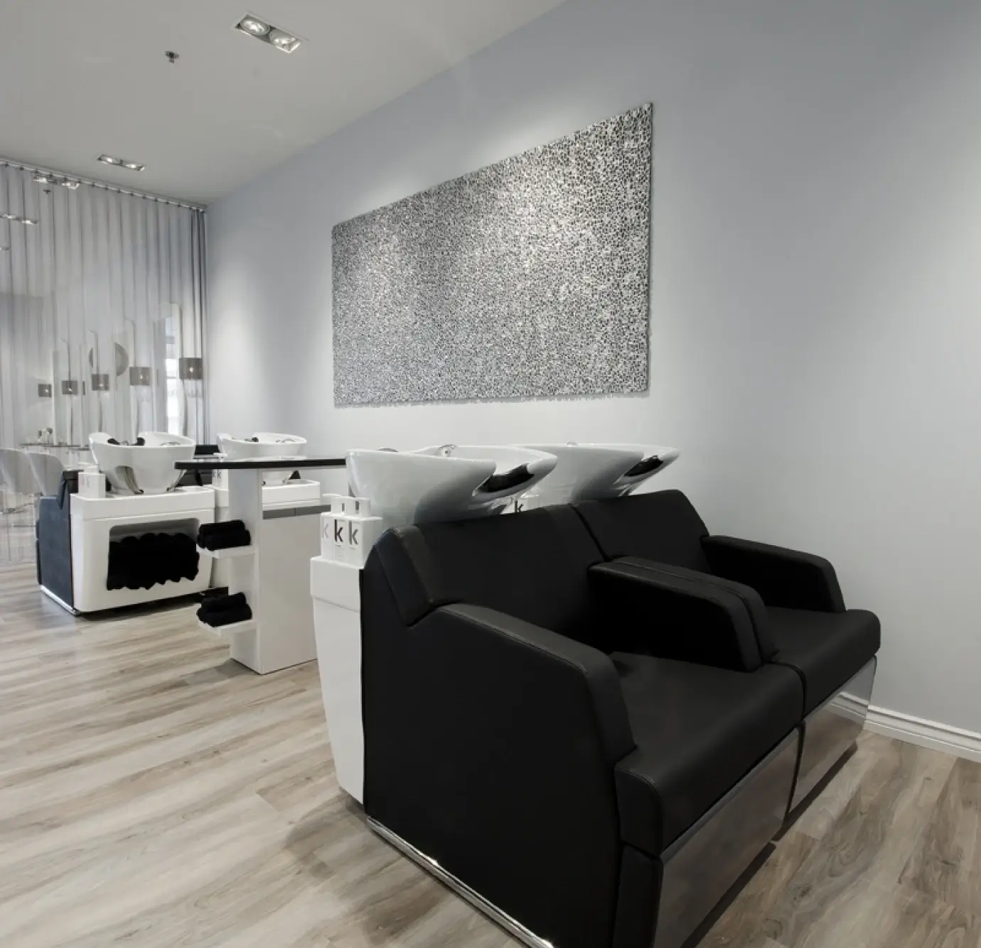 Contemporary black and white salon shampoo area with two black leather wash chairs, white ceramic bowls on white pedestals, and large silver mosaic wall art
