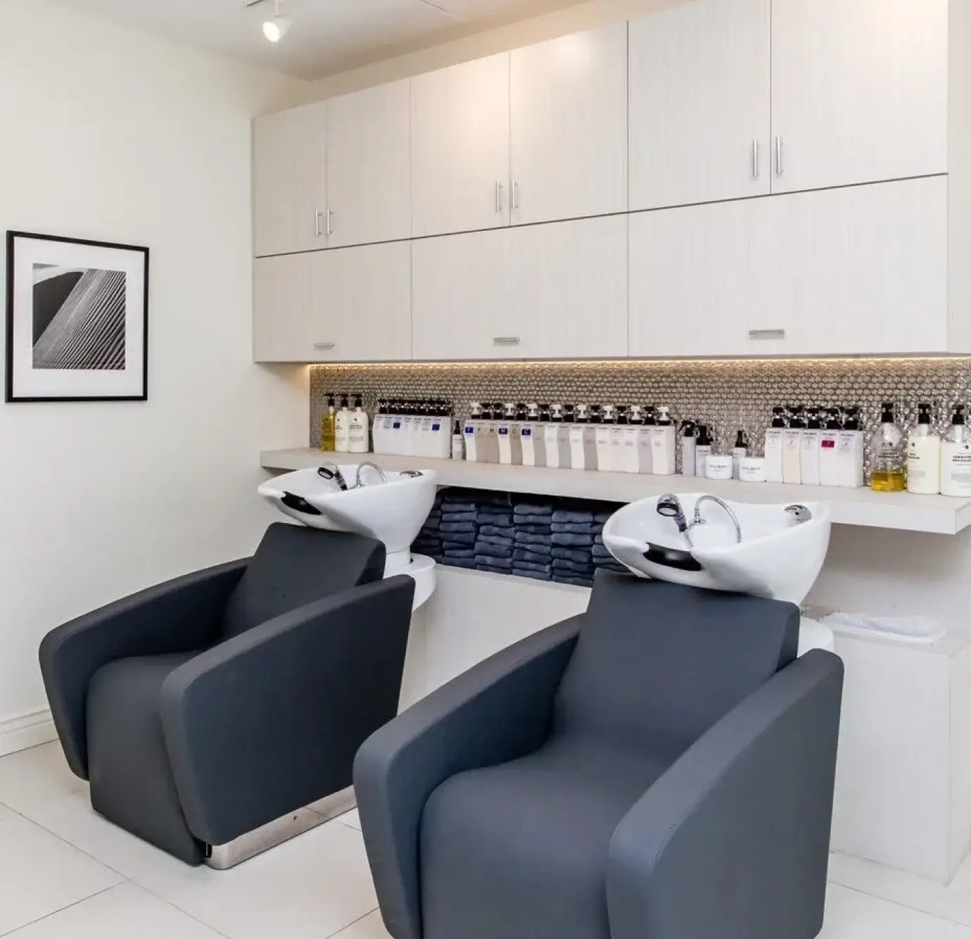 Clean minimalist salon shampoo room with two slate gray upholstered wash chairs, built-in white cabinetry, floating shelf with hair care products, and metallic penny tile backsplash