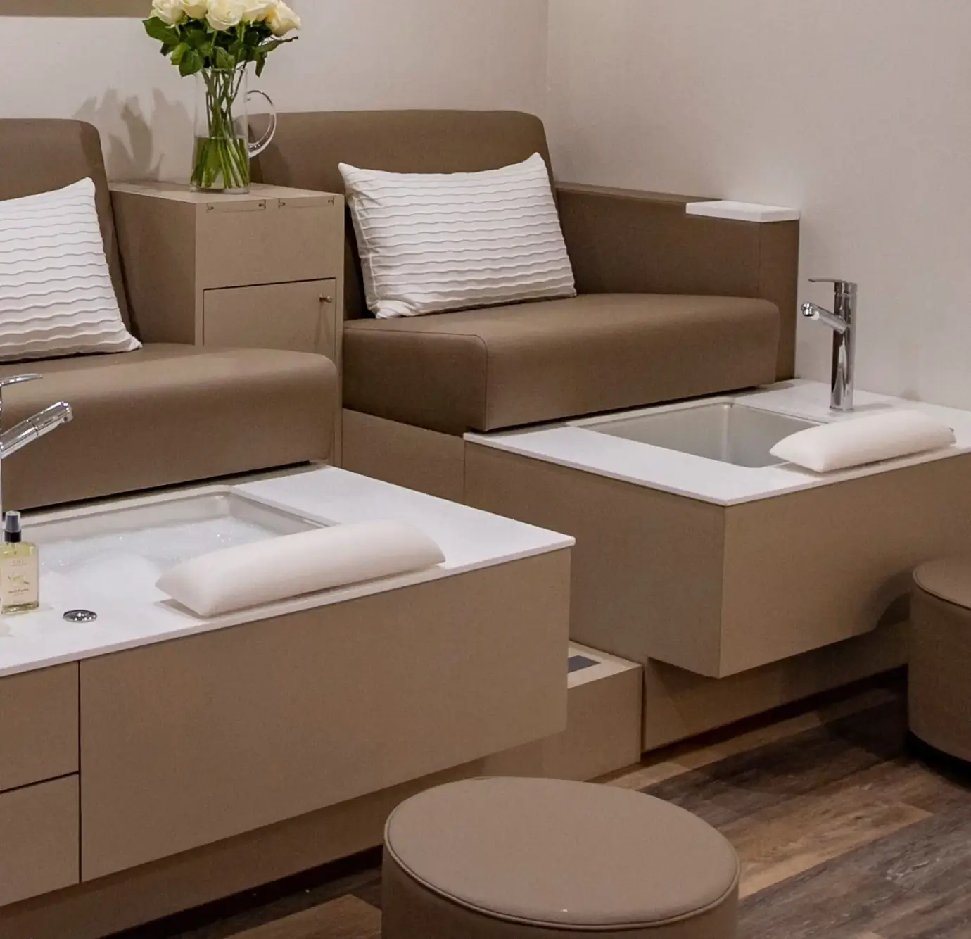 Contemporary spa pedicure chairs in taupe upholstery with white textured pillows, built-in foot soak basins with chrome faucets, matching ottoman stools, and white rose floral arrangement