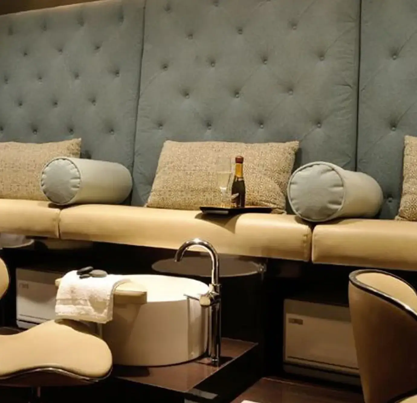 Glamorous nail salon pedicure lounge with floor-to-ceiling tufted seafoam blue upholstered wall, cream leather bench seating, gold shimmer lumbar pillows, cylindrical bolster pillows, and white porcelain foot basins with champagne service